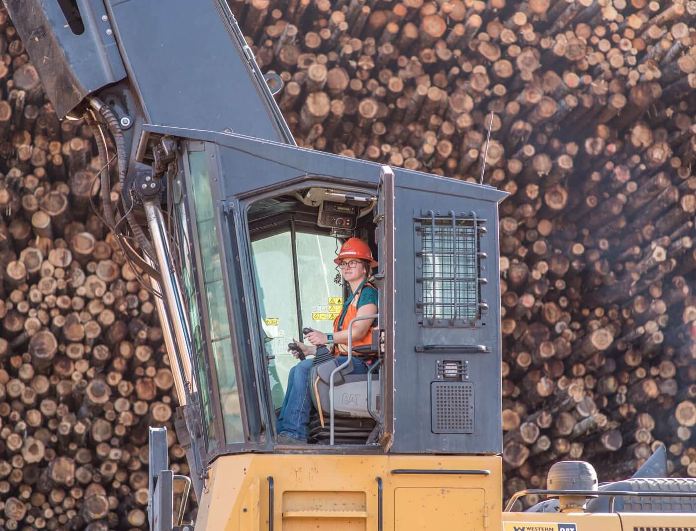A woman operating large machinery in front of a large pile of logs.