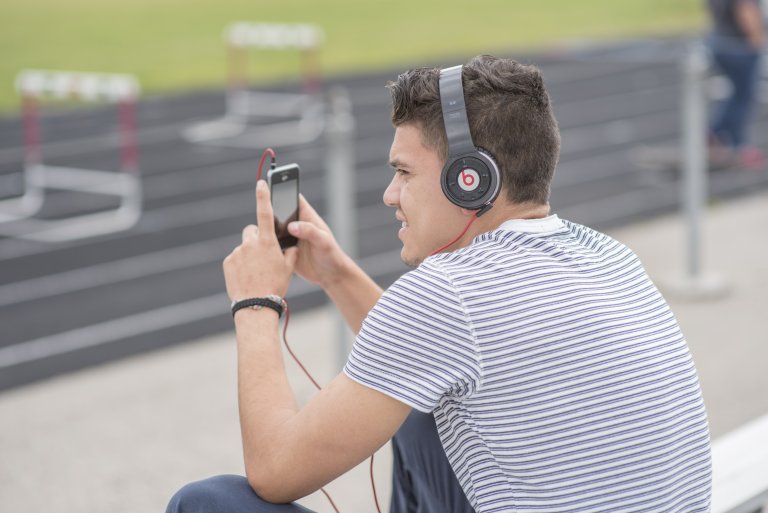 Student sitting beside a track looking at his phone and wearing headphones